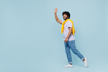 Full size body length side view young bearded Indian man 20s years old wears white t-shirt go meet greet waving hand as notices someone isolated on plain pastel light blue background studio portrait.