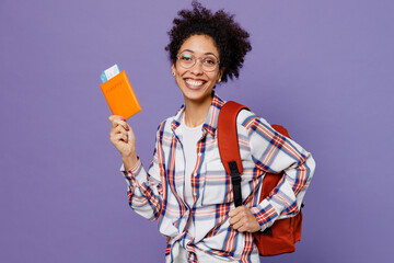 Young smiling tourist girl woman of African American ethnicity student in shirt backpack travel abroad hold passport ticket stand akimbo isolated on plain purple background Air flight journey concept