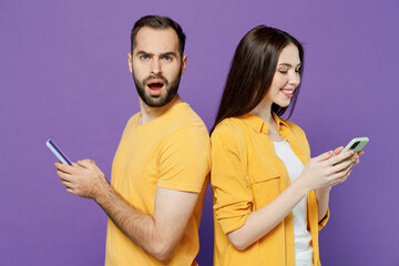 Side view young indignant jelaous couple two friends family man woman together in yellow clothes hold in hand use mobile cell phone stand back to back look camera isolated on plain violet background.