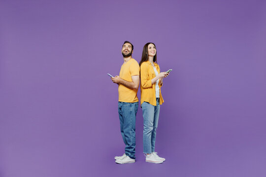 Full Body Side View Young Couple Two Friends Family Man Woman Together In Yellow Clothes Hold In Hand Use Mobile Cell Phone Stand Back To Back Browsing Internet Isolated On Plain Violet Background