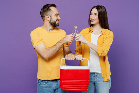 Young smiling fun couple two friends family man woman together wear yellow clothes hold red box freezer cooler refrigerator drink beer raise toast isolated on plain violet background studio portrait.