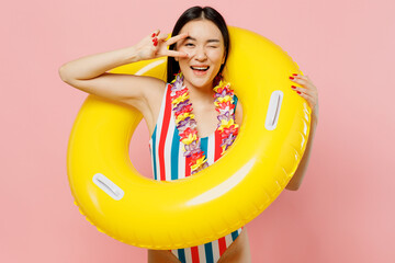 Young woman of Asian ethnicity in striped one-piece swimsuit hawaii lei look through inflatable ring cover eye with v-sign isolated on plain pink background. Summer vacation sea rest sun tan concept.