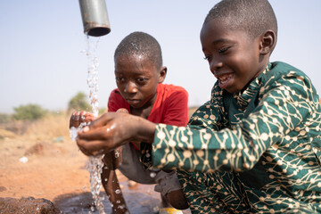 Two African boys enjoying the fresh water pouring from a tap at rural village well; water as basic...