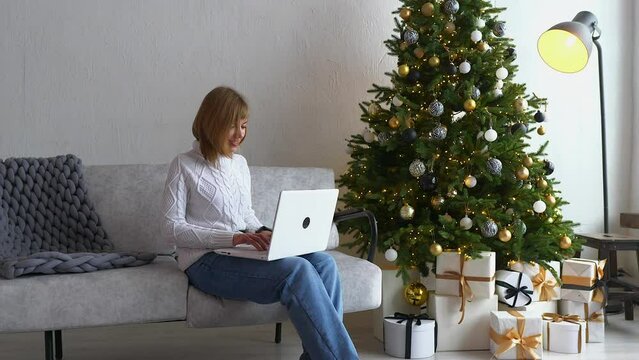 Beautiful woman is having a video call, online conversation on her laptop, while spending time at home at Christmas holidays. Happy girl is sitting on the sofa next to Christmas tree with presents