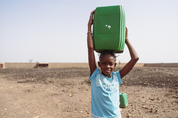 Arid African countryside with a young child lifting a heavy water canister on her head to provide...