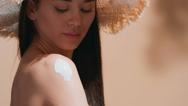 Close-up Beauty Portrait Of Dark-haired Girl In A Straw Hat Applying Sunscreen On Her Shoulder On Beige Background | Sunscreen Application Shot For Beauty Commercial