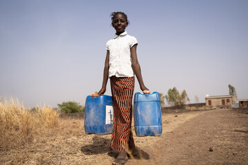 Skinny African girl carrying two water containers in the middle of an arid landscape; concept of poverty, lack of pipe connections in private rural homes, child labor; Millennium Development Goals - obrazy, fototapety, plakaty
