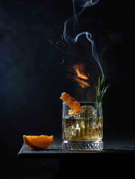 Whiskey or cognac in cristal glass with smoked rosemary,fire and slice of orange on black table.Close up of alcoholic drink.