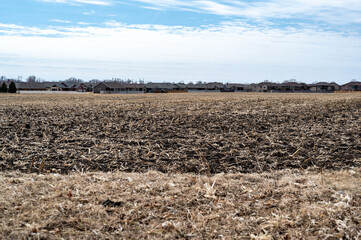 Empty corn field after fall harvest with residue over soil. Urban sprawl visible in the distance with residential housing development.