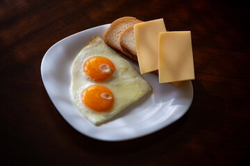 a plate with two eggs, fresh white bread and two slices of cheese in the sunlight on a dark wooden background
