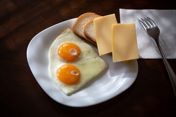 a plate with two eggs, fresh white bread and two slices of cheese in the sunlight on a dark wooden background