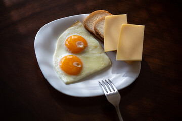 a plate with two eggs, fresh white bread and two slices of cheese in the sunlight on a dark wooden background