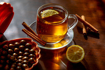 A cup of hot freshly brewed tea with lemon, cinnamon and chocolates in the sun on a dark wooden background