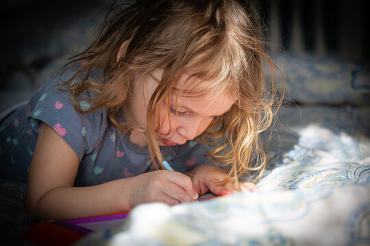 A little girl is lying on the bed and drawing on a graphic tablet in the sunlight
