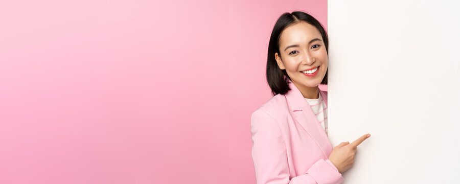 Image of asian female entrepreneur in suit, pointing finger at board, showing smth on white wall, demonstrating chart or information, standing over pink studio background