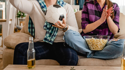 Close up on hands of unknown caucasian man holding soccer football ball while watching game at home with his girlfriend or wife couple