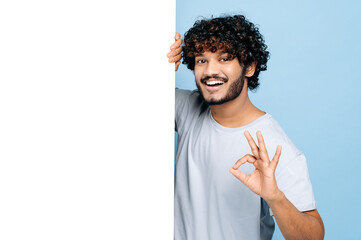 Happy Indian or Arabian guy, in casual t-shirt, peeking out from behind advertisement white board,...