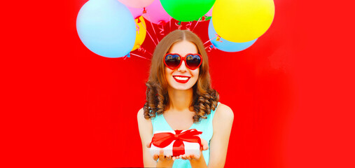 Portrait of happy smiling young woman with gift box and colorful balloons on red background