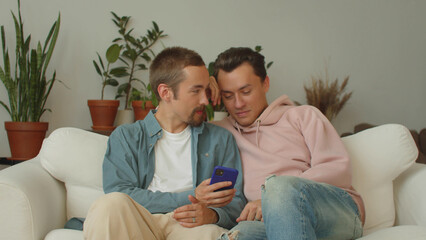 Lgbt men are sitting on the sofa in the living room and watching something on the phone in the Internet, then smiling