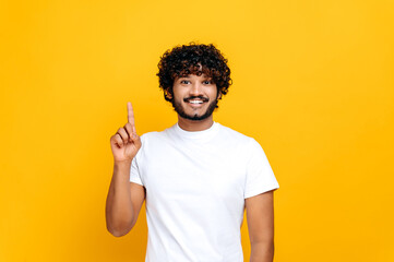 Joyful indian or arabian guy in basic white t-shirt, amazed looks at camera and points finger up, at empty copy space, stands on isolated orange color background, smiling. Mock-up, copy space concept