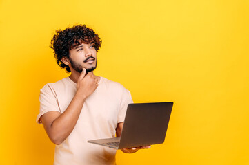 Handsome charismatic indian or arabian curly guy in basic t-shirt, holding open laptop in hand, looking away thoughtfully, planning, dreaming, standing over isolated orange background