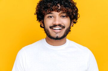 Close-up portrait of handsome attractive positive curly- haired indian or arabian guy, wearing white basic t-shirt, standing over isolated orange background, looking at camera, smiling friendly