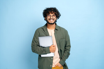 Smart handsome positive indian or arabian guy, with glasses, in casual wear, student or freelancer, holding a laptop in hand, standing on isolated blue background, looking at camera, smiling friendly - obrazy, fototapety, plakaty