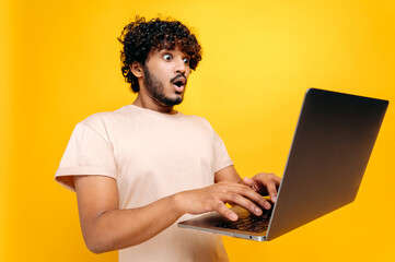 Excited shocked indian or arabian curly-haired guy, typing on a laptop, looking in surprise at the screen, saw unexpected news, victory, profit, stands on isolated orange background. Emotional face