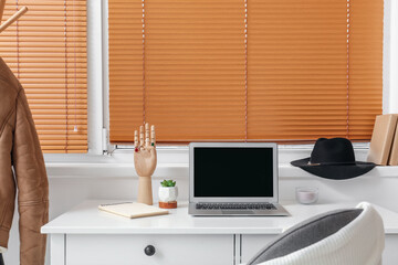 Modern laptop, wooden mannequin hand and houseplant on table near window