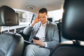 Young businessman tired from work holding hand to his head while sitting in the back seat of the car.