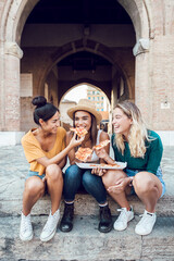 Vertical shot of happy multiracial women eating pizza in city street - Three diverse female tourist...