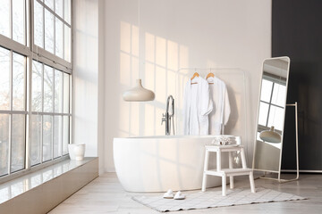 Interior of stylish bathroom with bathtub, mirror and rack with bathrobes near white wall