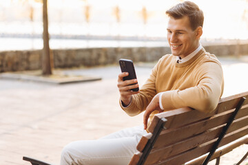 Modern handsome smiling man in yellow and white clothes talking on the phone sitting on a park bench at sunset