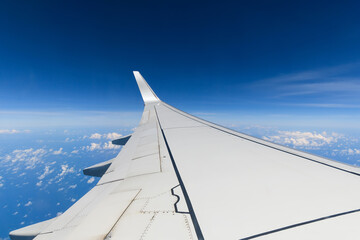 View of the wing of a white airplane during a flight on a beautiful blue sky day with few white clouds, flying high.
