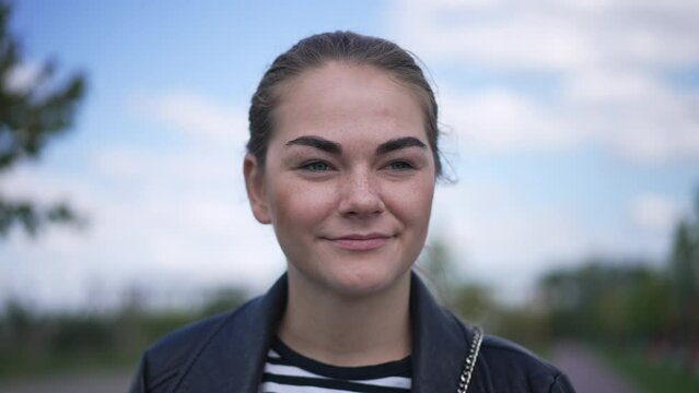 Headshot portrait of freckled beautiful Caucasian young woman looking away standing outdoors. Attractive confident millennial lady in summer park with clouds on sky at background. Slow motion