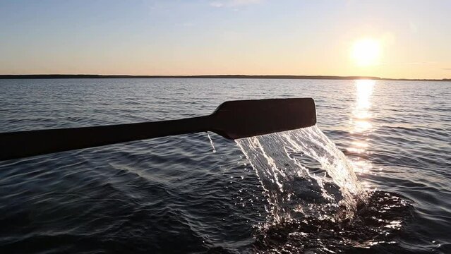 A person is rowing with a wooden oar on a boat on a lake during sunset. 