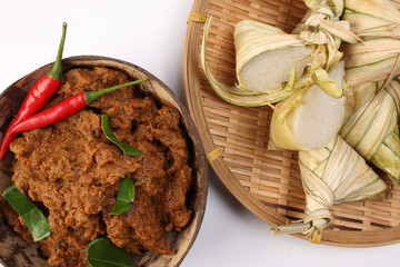 Traditional dry beef curry rendang in coconut shell with diamond shaped woven palm leaf pouch steamed glutenous rice cake ketupat kupat tipat on bamboo tray white background
