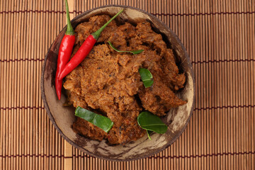 Traditional cooked dry curry beef meat rendang closeup in coconut shell bowl on bamboo mat rustic wooden background