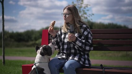 Wide shot woman eating croissant drinking coffee with curios dog watching owner. Portrait of young beautiful Caucasian lady enjoying morning with pet outdoors in city park. Slow motion