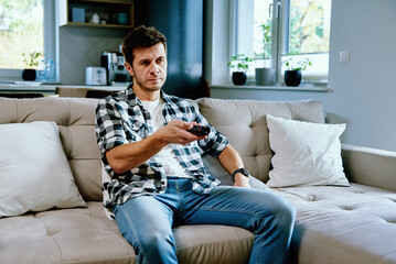 Obraz premium Man sitting on sofa and watching TV, switch channels using remote control. Resting at home, leisure weekend