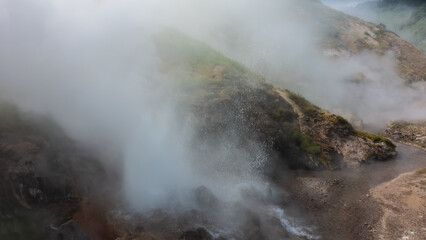 The eruption of a geyser on the hillside. Thick steam rises above the boiler, splashes of hot water. The river flows along a rocky bed. Kamchatka. Valley of Geysers