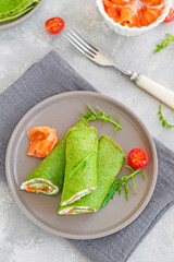 Green spinach crepes or pancakes with smoked salmon and soft cheese on a plate on the gray background. Copy space.