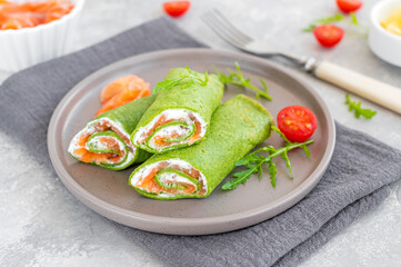 Green spinach crepes or pancakes with smoked salmon and soft cheese on a plate on the gray background. Copy space.