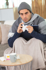 poorly man wearing warm clothing on sofa holding tissue