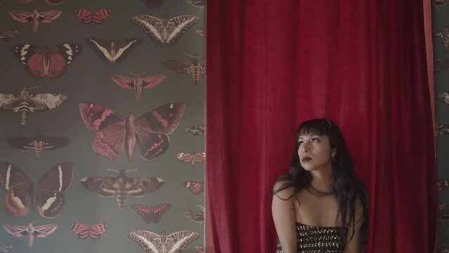 A tracking shot of a beautiful woman wearing a tube top while dancing sensually in front of a red curtain hanging on a wall of various butterfly photographs. Her body language is so sexy and tempting.