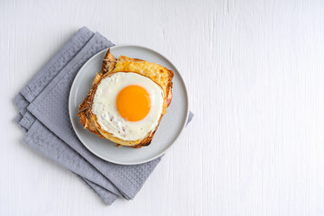 Top view of Croque madame french sandwich made of toasted bread, bechamel sauce, ham and cheese baked into a crunchy snack with fried egg served on plate on white wooden background with copy space