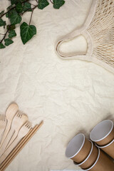 Disposable bamboo cutlery in disposable paper cups on canvas background and natural ivy branch. Flat lay, place for text. Vertical image.