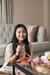 Cute asian girl sitting on floor in living room and painting picture with watercolor.