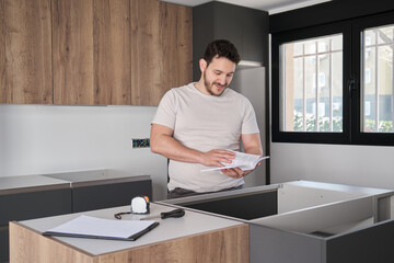 Young hispanic man reading instruction manual before installing furniture on modern new kitchen.