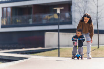 black-haired thin woman following a little African-American boy with curly hair standing on the scooter and looking sideways full shot urban background. High quality photo
